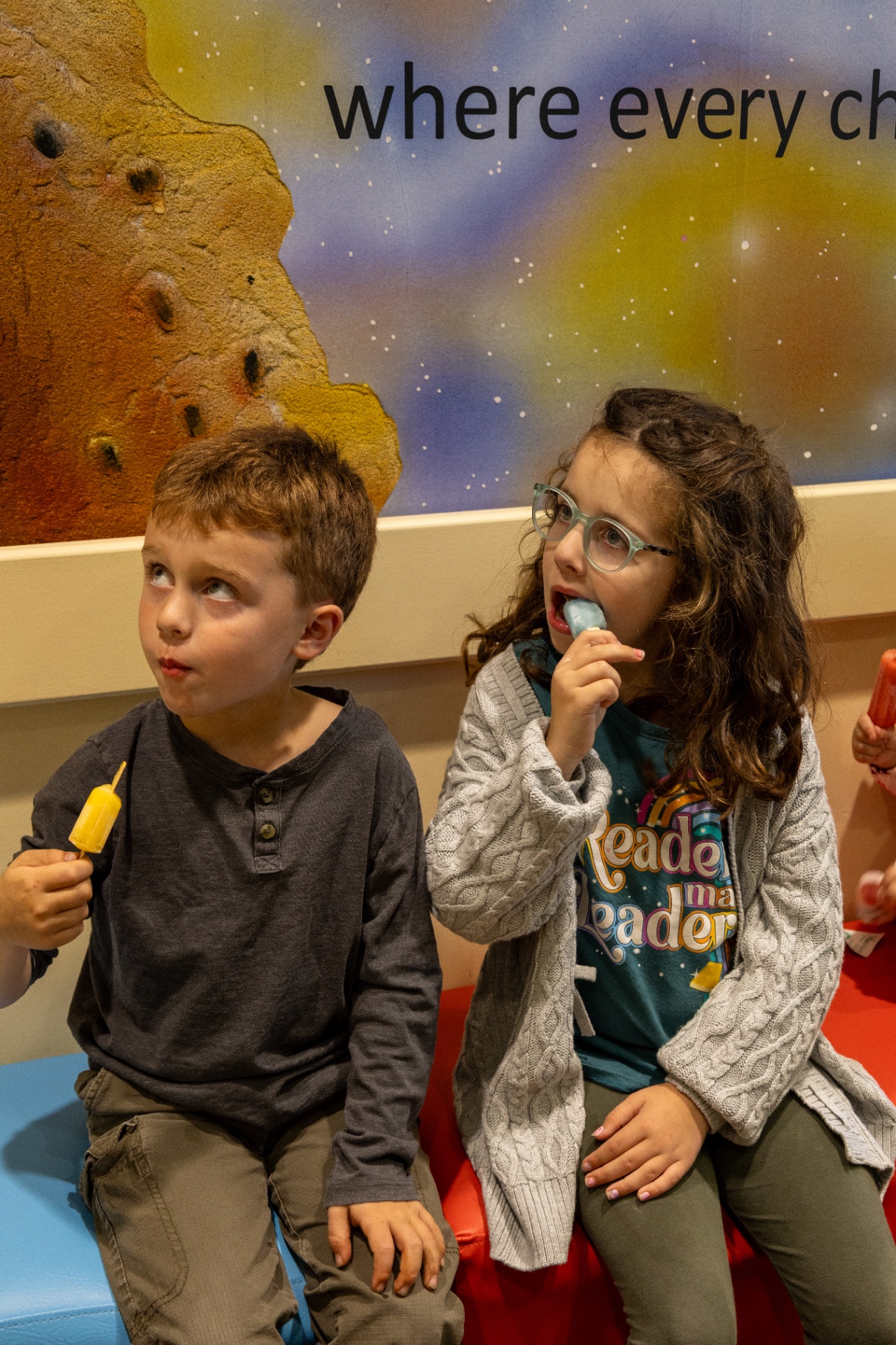 Two children eating freezies on a bench beneath the waiting room space mural
