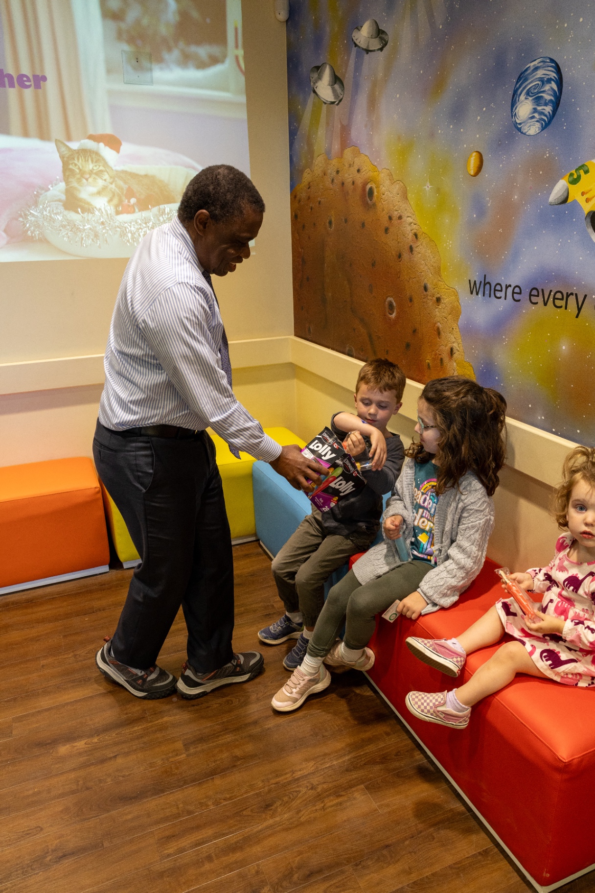 Dr Ukpeh handing out freezies to children seated on a red bench in the waiting room