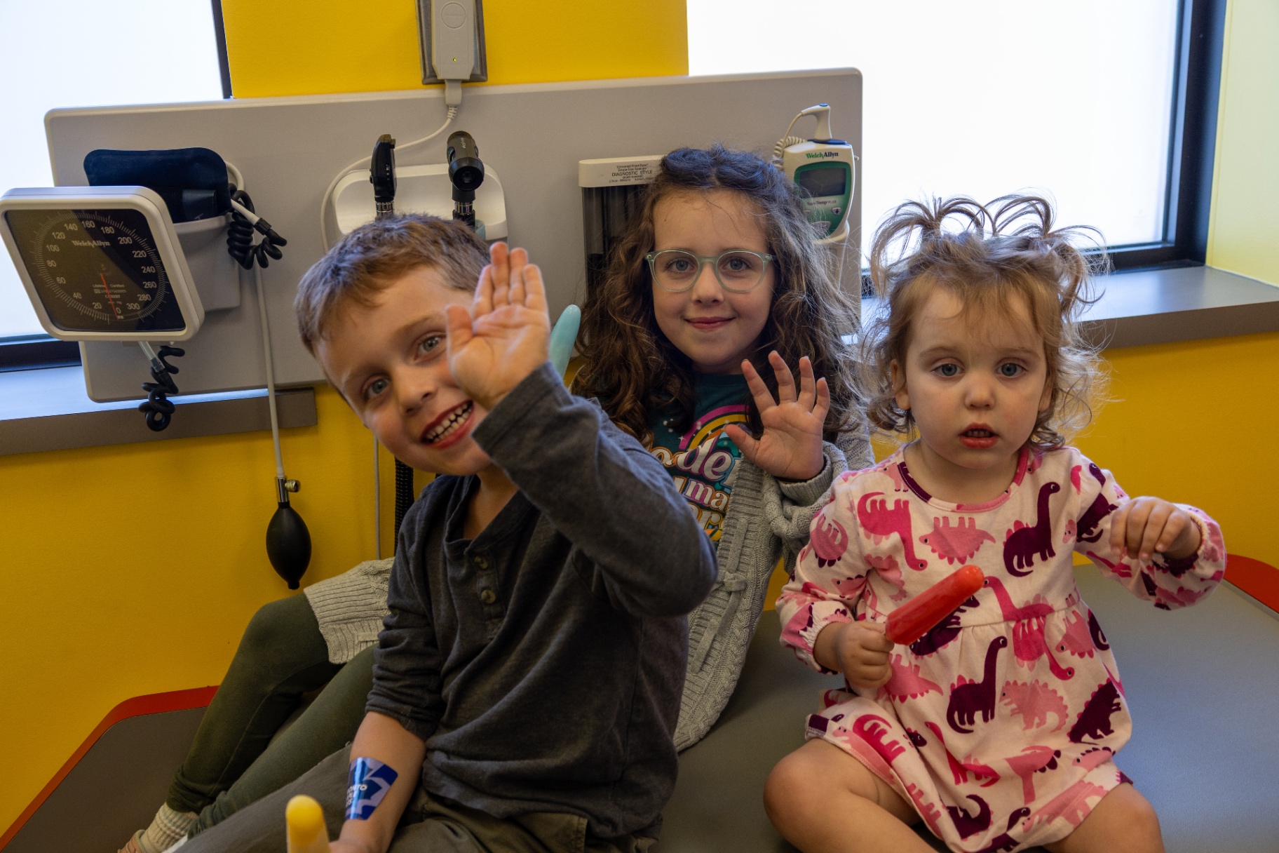Three children sitting on an exam table waving at the camera