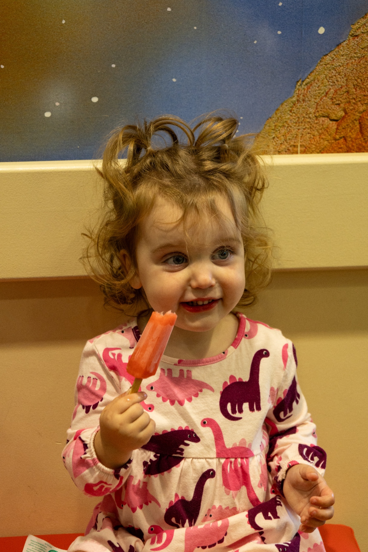 Young girl in a dinosaur print dress smiling and holding an orange freezie